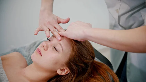 Massaggio - massaggiatrice massaggiatrice che massaggia il viso di una donna dai capelli rossi con le mani — Foto Stock