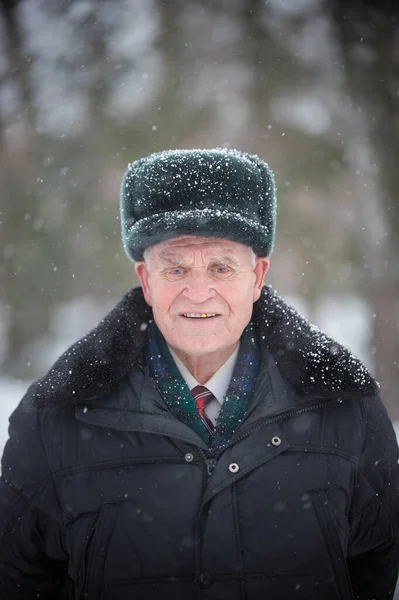 An old smiling man in warm hat standing outdoors at winter and looking in the camera