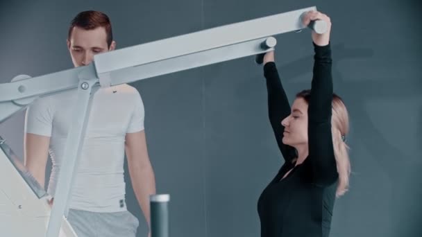 Entrenamiento con instructor de fitness en el gimnasio moderno - una joven entrenando sus manos en el simulador moderno — Vídeos de Stock