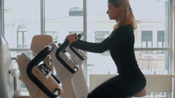 Una joven vestida de negro haciendo ejercicios en sus piernas en bicicleta estática en un gimnasio blanco — Vídeos de Stock