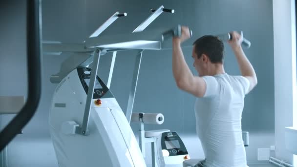 Moderní tělocvična - muž sedící na tréninkovém přístroji a cvičící si ruce v bílé moderní tělocvičně — Stock video