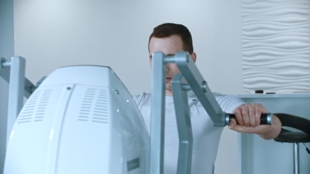 现代体育馆-一个穿着白色T恤的年轻英俊男子在训练器械上做手臂运动 — 图库视频影像