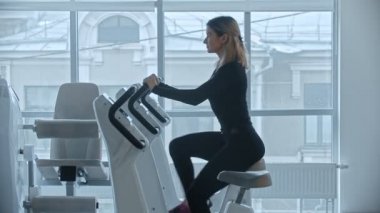 Siyah elbiseli genç bir kadın spor salonunda egzersiz bisikletine binip büyük aynaların arkasına oturur.