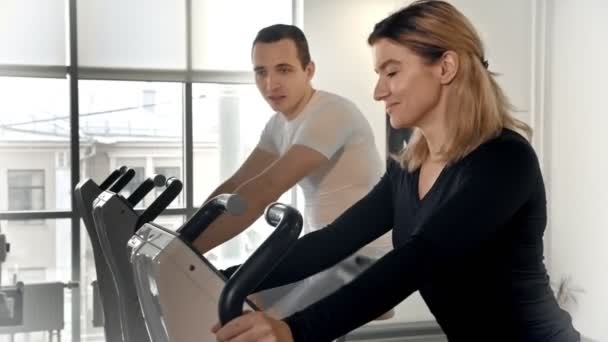 Sportif erkek ve kadın bacaklarını egzersiz bisikletinde çalıştırıyor ve birbirleriyle konuşuyorlar. — Stok video