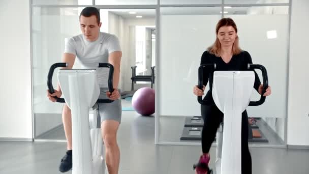 Deportivo sonriente hombre y mujer entrenando sus piernas en bicicleta estática y hablando entre sí — Vídeos de Stock