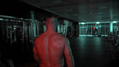 Yetişkin, üstsüz, vücut geliştiren bir adam karanlık bir spor salonunda neon ışıkla yürüyor.