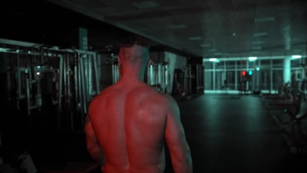 Un bodybuilder adulto senza maglietta che cammina nella palestra buia con illuminazione al neon — Video Stock