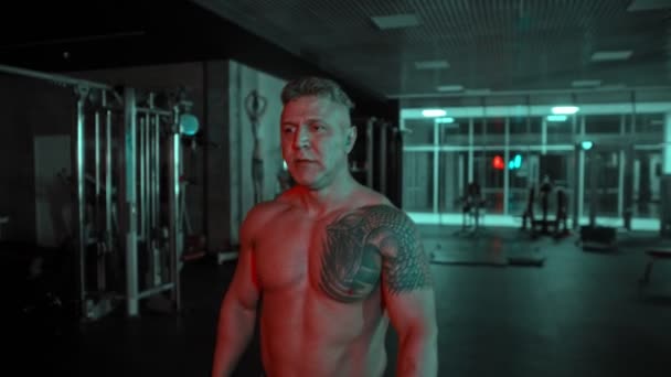 Een volwassen shirtloze man bodybuilder wandelen in de donkere sportschool met neon verlichting en kauwgom — Stockvideo