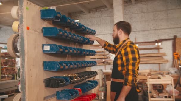 Trabajador barbudo eligiendo tornillos desde el stand en el taller de carpintería y se aleja — Vídeo de stock