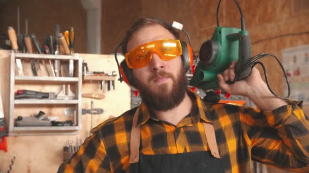 Trabajador barbudo en camisa amarilla y gafas protectoras de pie en el taller sosteniendo el instrumento de molienda — Vídeos de Stock