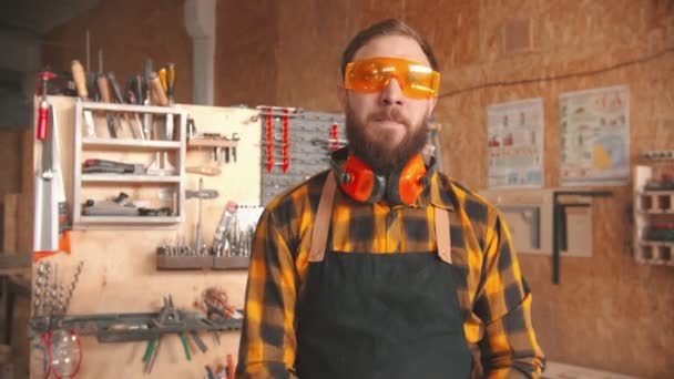 Trabajador barbudo con camisa amarilla y gafas protectoras parado en el taller y poniéndose los auriculares — Vídeos de Stock