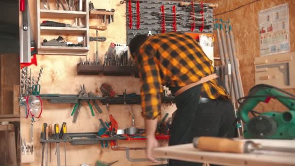 Trabajador barbudo en camisa amarilla eligiendo instrumentos en el taller y toma la rectificadora de la mesa — Vídeo de stock