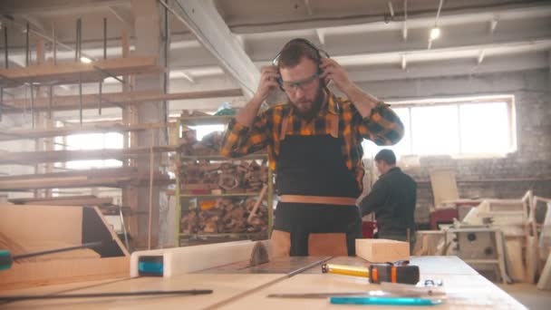 Hombre barbudo poniéndose auriculares y comienza a cortar la madera contrachapada — Vídeos de Stock