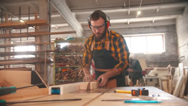 Γενειοφόρος άνθρωπος που βάζει ακουστικά και αρχίζει να κόβει ένα κομμάτι ξύλο με ένα δισκοπρίονο — Αρχείο Βίντεο