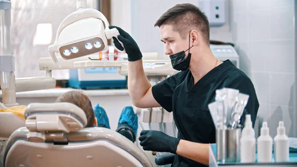 Homem dentista abaixa a lâmpada para baixo para examinar a boca de um menino — Fotografia de Stock