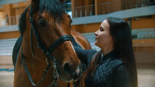 Ипподром - молодая улыбающаяся женщина в специальном снаряжении стоит с лошадью и гладит ее — стоковое фото