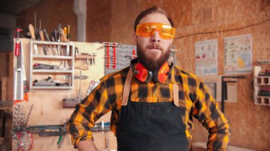 Sarı gömlekli ve koruyucu gözlüklü sakallı adam atölyede duruyor. Eller belde.