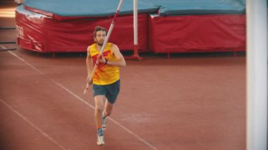 Stadyumda sırıkla atlama eğitimi alan atletik bir adam barın üzerinden atlıyor.