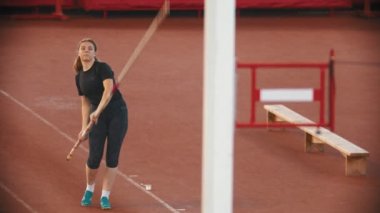 Stadyumda sırıkla atlama eğitimi alan bir kadın barın üzerinden atlayıp ona dokunuyor.