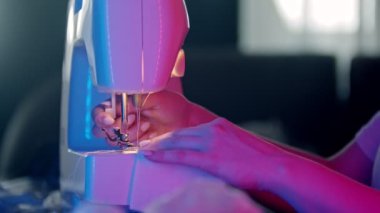 Genç kadın terzinin elleri, beyaz bir bez dikiyor, neon ışıkta dikiş makinesi kullanıyor.
