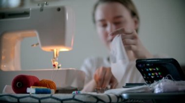 Genç bir kadın dikiş makinesi kullanarak beyaz koruyucu bir maske dikiyor.