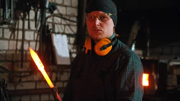 Forging industry - a man blacksmith holding a hot piece of metal and looking in the camera