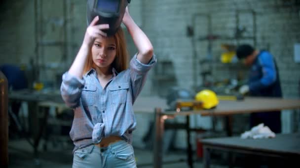 Young sexy woman in small shorts standing in the workshop and putting on her helmet - man working on the background — Stock Video