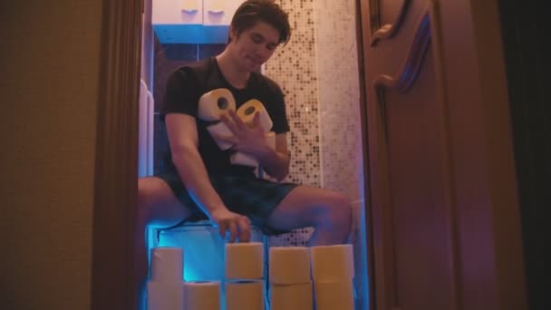 Een jongeman die op een toilet zit met een hoop toiletpapier en er een muur van maakt. — Stockvideo