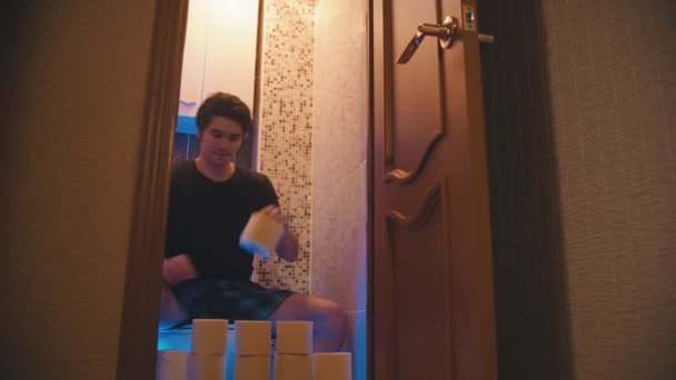 Молодой человек сидит на унитазе с кучей туалетной бумаги и закрывает дверь — стоковое видео