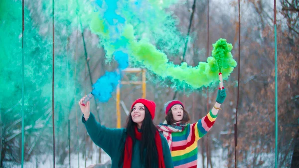 Twee jonge vrouwen op de besneeuwde brug hebben plezier met groene en blauwe rookbommen — Stockfoto