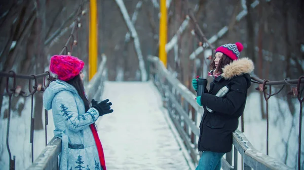 Duas jovens mulheres bebem bebidas quentes da garrafa térmica e caminham na ponte — Fotografia de Stock