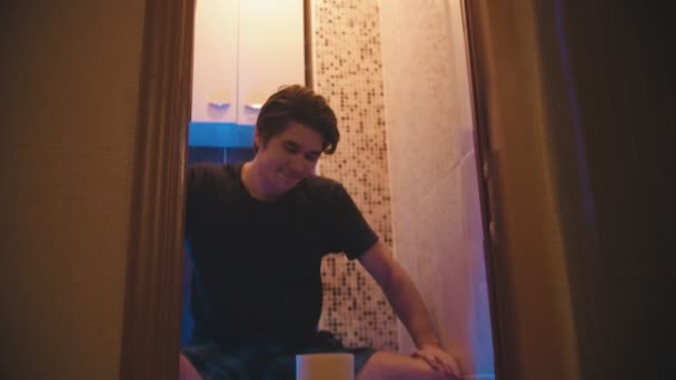 Молодий чоловік відкриває двері, сидячи на туалеті з купою туалетного паперу і посміхаючись — стокове відео