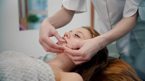 Massaggio - massaggiatrice massaggiatrice che massaggia il viso di una donna dai capelli rossi con le mani — Foto Stock