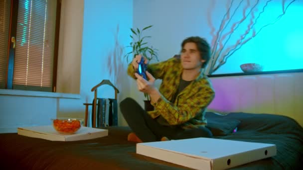 Νεαρός ενθουσιασμένος άντρας κάθεται στο κρεβάτι παίζοντας βιντεοπαιχνίδι και τρώγοντας πατατάκια — Αρχείο Βίντεο