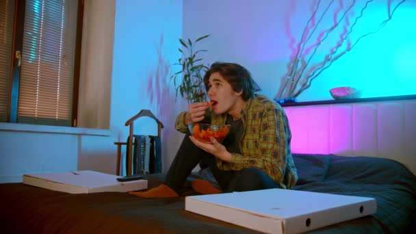 Joven sentado en la cama viendo televisión y comiendo papas fritas — Vídeos de Stock