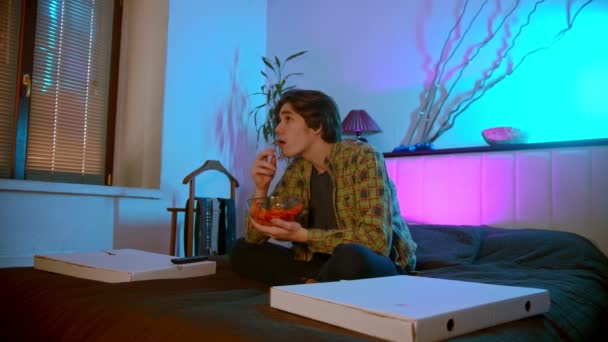 Jongeman zit op het bed tv te kijken en chips en andere snacks te eten — Stockvideo