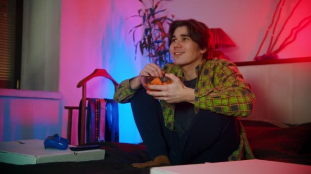 Young man sitting on the bed watching movie and eating chips — Stock Video