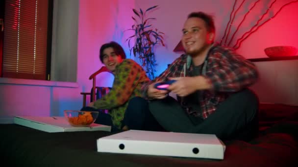 Två unga män vänner sitter på sängen och spela spel med gamepads och ha kul — Stockvideo