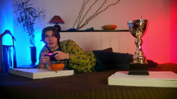 Νεαρός άνδρας με ακουστικά ξαπλωμένος στο κρεβάτι παίζοντας online βιντεοπαιχνίδι — Αρχείο Βίντεο