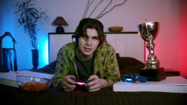 Kulaklıklı genç adam yatakta uzanıyor, internette video oyunu oynuyor ve mikrofonla konuşuyor. — Stok video