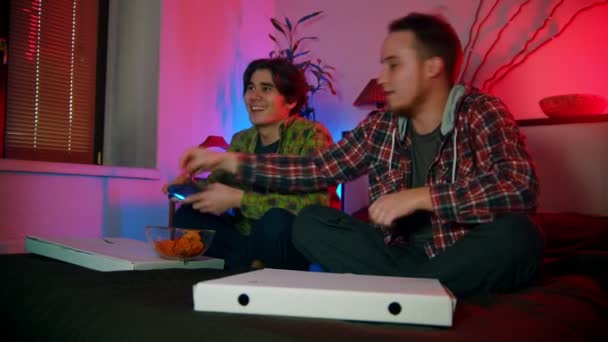 Dva mladí muži přátelé jíst chipsy a hrát hru s gamepady - jeden z nich prohrává — Stock video