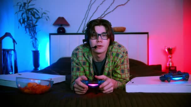 Νεαρός όμορφος άντρας με γυαλιά και ακουστικά ξαπλωμένος στο κρεβάτι παίζοντας online βιντεοπαιχνίδι — Αρχείο Βίντεο