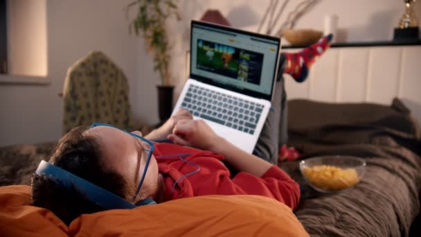 Молодой человек лежит на кровати и смотрит видео с ноутбука и ест чипсы — стоковое видео