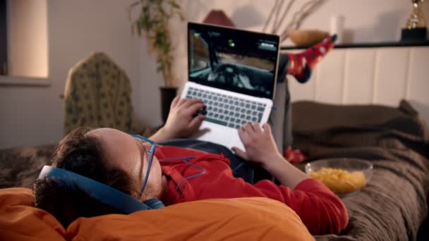 Молодой человек лежит на кровати и играет в видеоигры на ноутбуке — стоковое видео