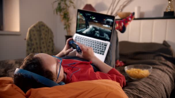 Молодой человек, лежащий на кровати, играет в гоночную видеоигру на ноутбуке с геймпадом — стоковое видео