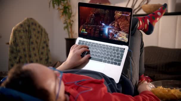 Молодой человек лежит на кровати и играет в видеоигру на ноутбуке — стоковое видео