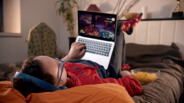 Молодой человек лежит на кровати и играет в шутер на ноутбуке — стоковое видео