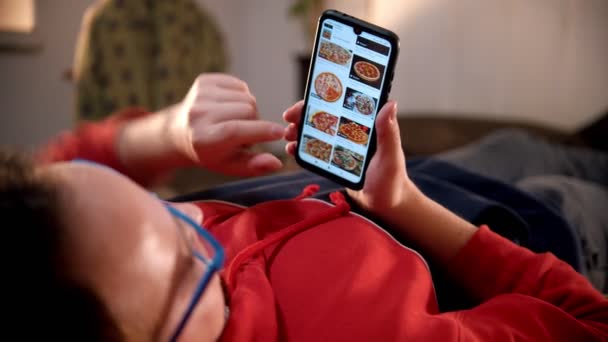 Joven acostado en la cama y desplazando fotos de pizza en su teléfono — Vídeo de stock