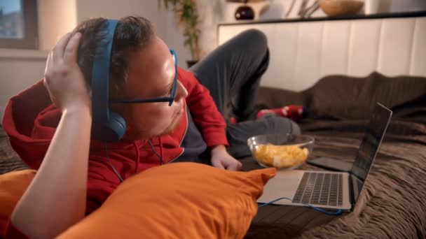 Joven con auriculares acostado en la cama y viendo películas — Vídeos de Stock