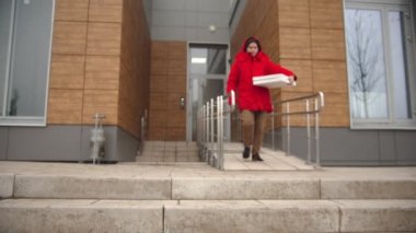 Kırmızı elbiseli bir kurye binadan çıkıyor ve pizza kutusundan bir dilim pizza alıyor.